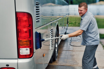 driver washes large bus with brush in open parking lot on summer day. Caucasian man prepares bus to...