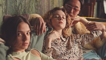 Two attractive girls and pompous Asian guy in fur coat looking at camera and making seductive moves sitting on sofa in authentic studio