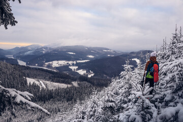 Candid portrait of young smiling man wearing colourful winter hiking jacket, warm-ups and white cap overlooking the snowy area of Stare hamry with the highest point of the Beskydy Mountains Lysa hora