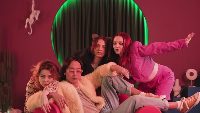 Slowmo of three expressive girls performing eccentric vogue moves while dancing to pompous confident Asian boy in glamorous outfit sitting on chair and looking at camera