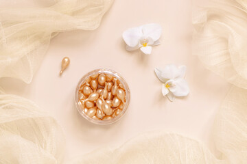 Face serum capsules in a jar near orchid flowers on light beige top view. Beauty product