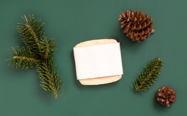 Soap bar with blank label near fir tree branches and pine cones on green top view, cosmetic mockup