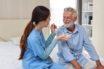 Obraz premium Happy caregiver sitting and feeding senior caucasian man on bed with rice porridge soup for breakfast at retirement house. Asian smiling nurse taking good help care and support elder patient at home.