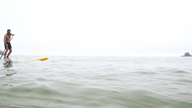 An athletic man paddles intensively on a large yellow SUP and swims through the sea waves. Extreme outdoor recreation.