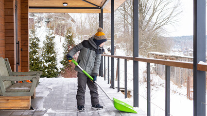 Involving children in housework concept. The boy cleans the snow on the terrace of his house with a...