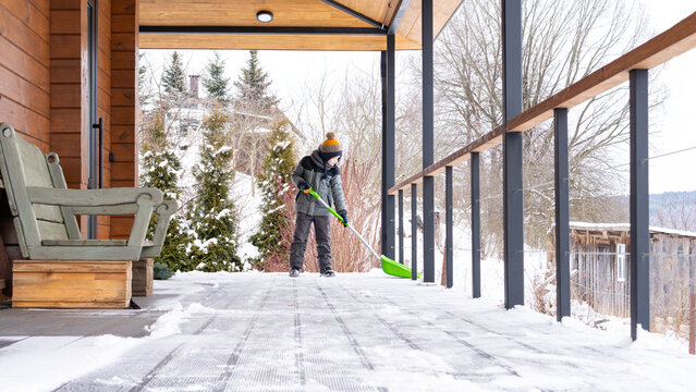 The Boy Cleans The Snow On The Terrace Of His House With A Big Shovel. Family Lifestyle In Winter. The Child Is Doing Household Chores. Kid Shoveling Snow On Sunny Day.