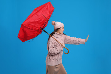 Young woman with umbrella caught in gust of wind on light blue background