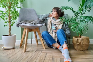 Cold winter season, woman with cat on chair near heating radiator