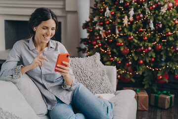 Happy female in earbuds holding smartphone, talking with family or friends online at Christmas time
