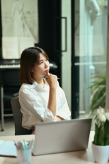 Thoughtful woman entrepreneur sitting at her workplace and looking through window, dreaming of success thinking of future business