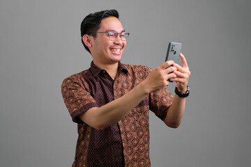 Cheerful young Asian man wearing batik shirt looking at smartphone, reading online news isolated on grey background