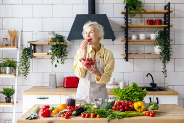 beautiful elderly gray haired senior woman cook in cozy kitchen with fresh organic vegetables, tomatoes, cabbage, lettuce, cucumbers on table cooking healthy vegetable salad, healthy food active life