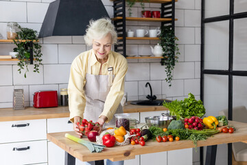 beautiful elderly gray haired senior woman cook in cozy kitchen with fresh organic vegetables, tomatoes, cabbage, lettuce, cucumbers on table cooking healthy vegetable salad, healthy food active life