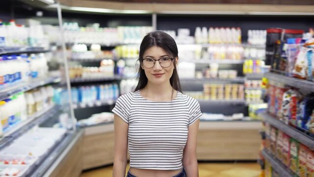 Portrait of attractive young asian woman standing in supermarket with shelves of dairy on background, looking at camera and smiling. Trade business and people concept