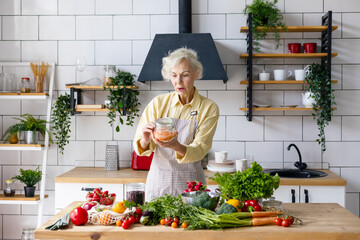 beautiful elderly gray haired senior woman cook in cozy kitchen with fresh organic vegetables, tomatoes, cabbage, lettuce, cucumbers on table cooking healthy vegetable salad, healthy food active life