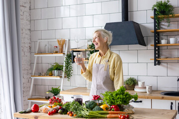 beautiful elderly gray haired senior woman cook in cozy kitchen with fresh organic vegetables on table for healthy vegetable salad, drink pure clear water from glass , healthy food for active life