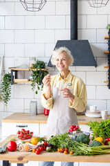 beautiful elderly grey haired senior woman in cozy kitchen takes vitamin supplement in capsule,...