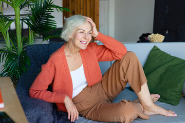Obraz premium happy beautiful elderly gray haired senior woman sit on sofa in living room in cosy apartment, smiling elderly woman, modern granny candid portrait