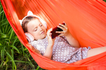 Cute little girl lies in a hammock with smartphone and wireless headphones and listening to music.