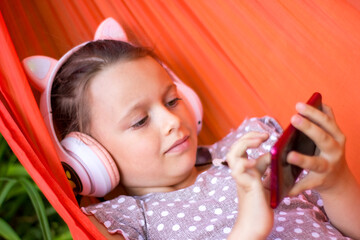 Cute little girl lies in a hammock with smartphone and wireless headphones and listening to music.