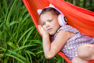 Cute little girl lies in a hammock and listening to music wearing wireless headphones at sunny summer day.