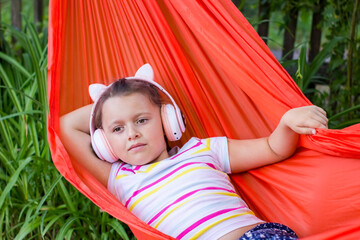 Cute little girl lies in a hammock and listening to music wearing wireless headphones at sunny summer day.