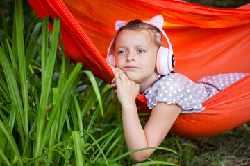 Cute little girl lies in a hammock and listening to music wearing wireless headphones at sunny summer day.