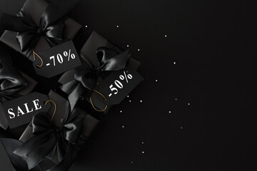 Top view photo of black gift boxes with black satin ribbons and sequins with text sale on price tag on black background, copy space. Black Friday Sale template.