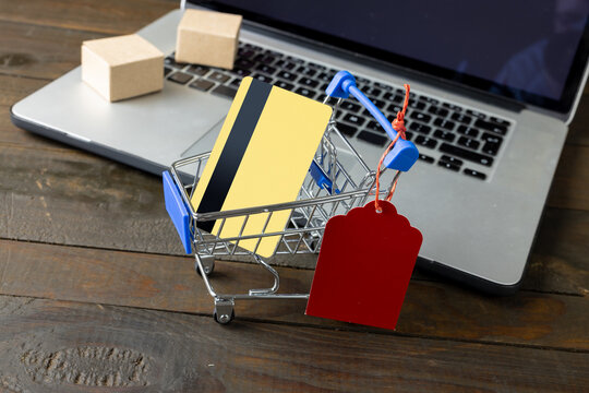 Composition of shopping cart with credit card and laptop on wooden background
