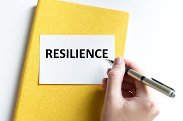 RESILIENCE text on a white card against the background of a yellow notebook next to the hand of the businessman with a pen writes the text