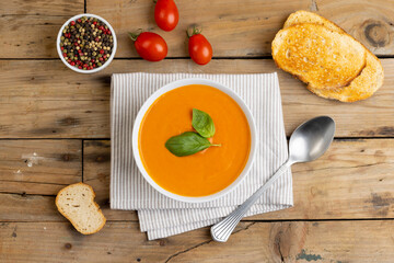 Horizontal image of bowl of tomato soup with basil garnish, tomatoes, toast and peppercorns on wood