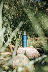 Vacuum thermos, camping flask on a stone by a stream in the forest on a sunny day. Equipment for hiking, camping