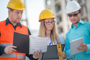 men and a woman in yellow and white helmets are standing at a construction site with documents and a tablet and discussing . The girl has a disgruntled face.
