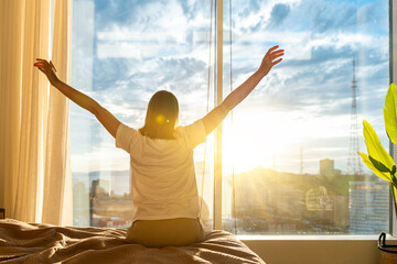 Fototapeta na wymiar Rear view woman sitting on bed in bedroom, stretching hands after awakening, dreamy young female enjoying morning, starting new day in modern apartment with large panoramic windows