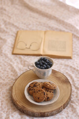 Vintage cup with fresh blueberries, plate of chocolate chip cookies, open book and reading glasses on a bed. Selective focus.