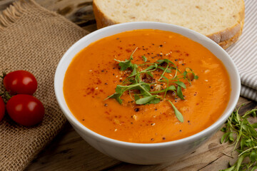 Horizontal image of bowl of tomato soup with garnish, tomatoes and sliced bread on wood