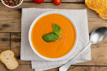 Horizontal image of bowl of tomato soup with basil garnish, tomatoes, toast and peppercorns on wood