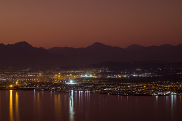 Landscape of sea, sea shore with city lights, mountains, cloudless sky at sunset and horizon