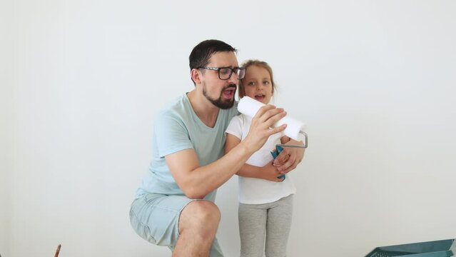 Happy family, young caucasian father and his cute child daughter singing and paints the wall with white paint. Repair in the apartment concept.