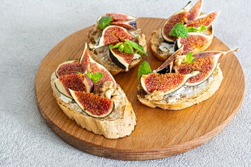 Crispy bruschetta (toast) with soft ricotta, ripe figs, walnuts and pine nuts, mint and honey on a wooden board on a light background.