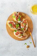 Crispy bruschetta (toast) with soft ricotta, ripe figs, walnuts and pine nuts, mint and honey on a wooden board on a light background.