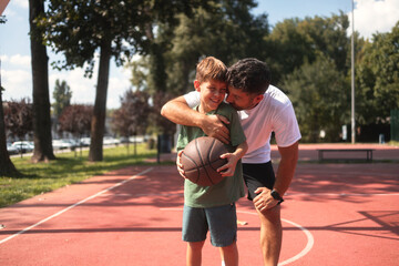 Smiling father hugging his son on basketball court. They wear casual clothes, boy holding a...
