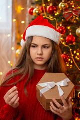 Unhappy girl with ended sparklers standing near Christmas tree with present box in her hands. Unpleasant moments in happy holidays times