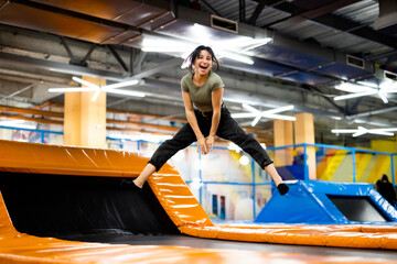 Naklejka premium Pretty girl jumping on colorful trampoline at playground park, posing and laughing. Beautiful female teenager happy during active entertaiments indoor