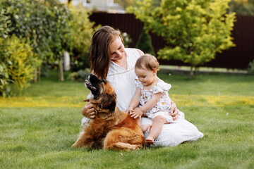 Family day, mother's day. Beautiful smiling young mom and baby daughter cuddling happy domestic dog on the backyard lawn.Mother with child girl are having fun with pet outdoors on summer holiday.