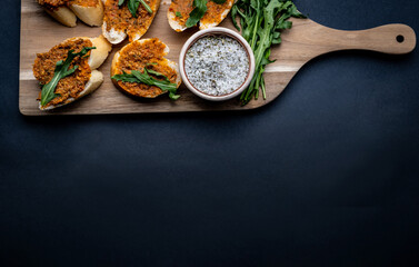 Mediterranean bruschettas with pesto and arugula served on wooden board with herbal salt and fabric on background with copy space. Traditional italian toasted bread with sauce and plants antipasti