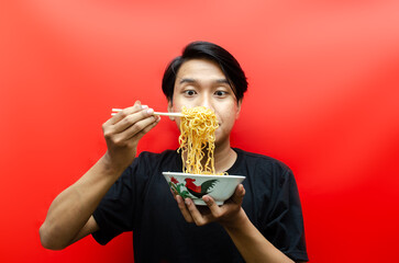 Portrait of Happy Asian man in black t-shirt eats instant noodles using chopsticks and bowl isolated on red background.