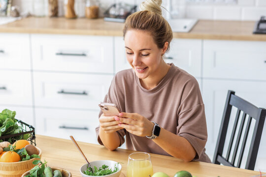 Healthy lifestyle concept. Young happy woman eating healthy food breakfast communicates in social media using mobile phone sitting at home in the kitchen in the morning