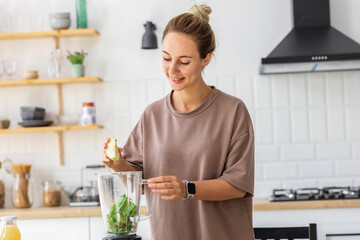 Caucasian female preparing breakfast from fresh green smoothie in the kitchen. Young happy woman cooking healthy food. Vegetarian food