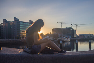Young girl with long hair sitting on the breakwater in the harbor reading a book in the light of the rising sun, golden hour photo shoot, Dublin, Ireland - obrazy, fototapety, plakaty